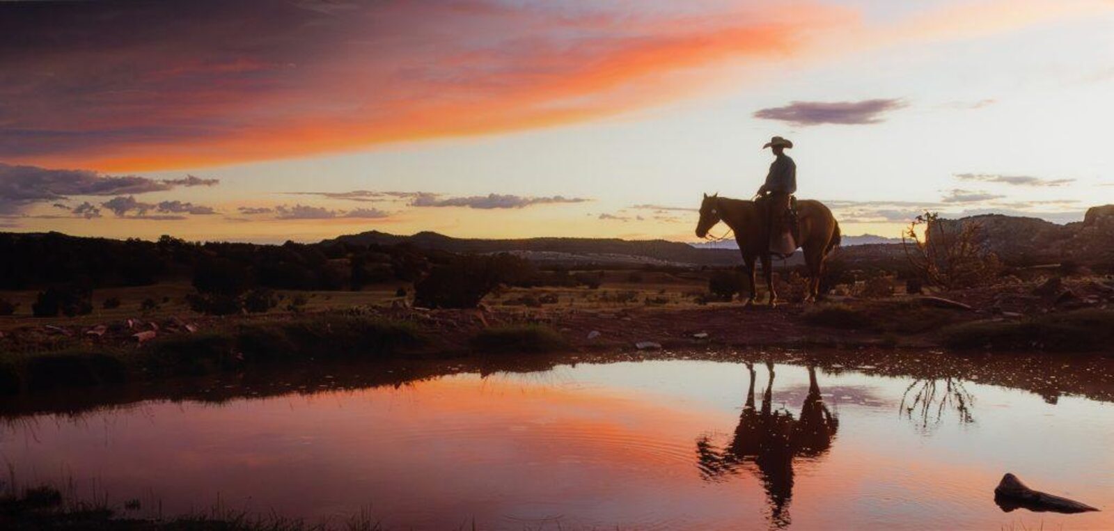 A cowboy on horseback at sunset reflected in a pond